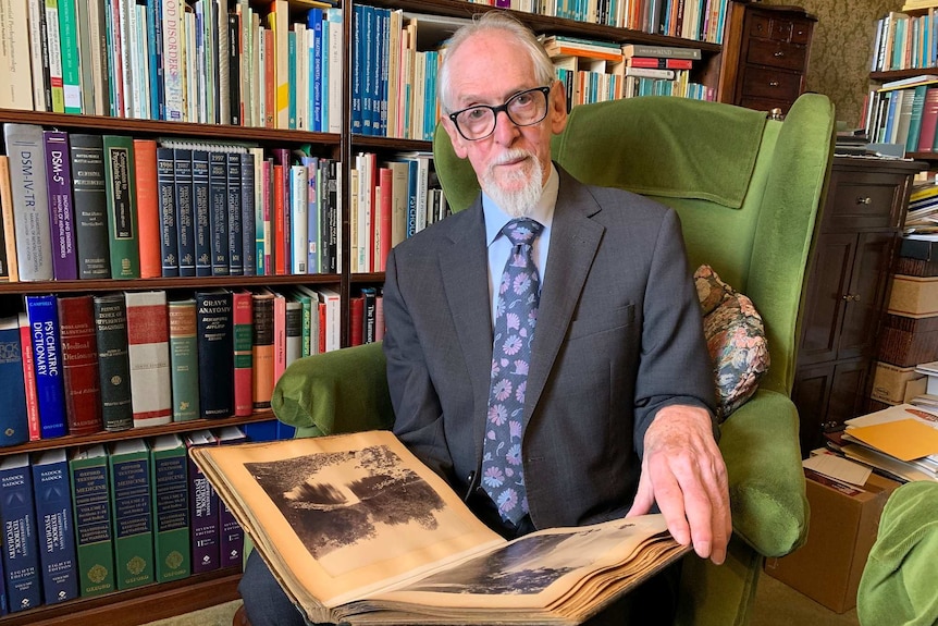 An older man sits in a green chair in front of shelves of books.