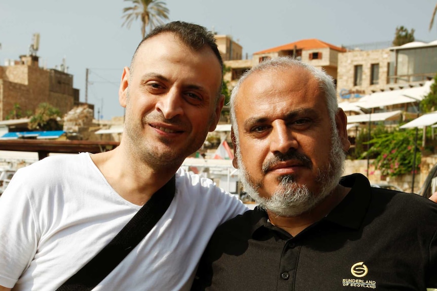Amer Khayat, in a white t-shirt with a shoulder bag, has his arm around the shoulder of Fadi, wearing a black polo shirt.