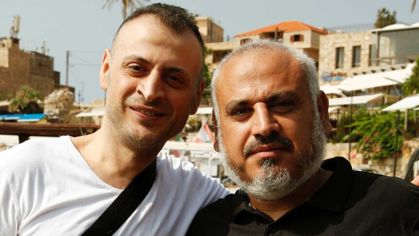 Amer Khayat, in a white t-shirt with a shoulder bag, has his arm around the shoulder of Fadi, wearing a black polo shirt.