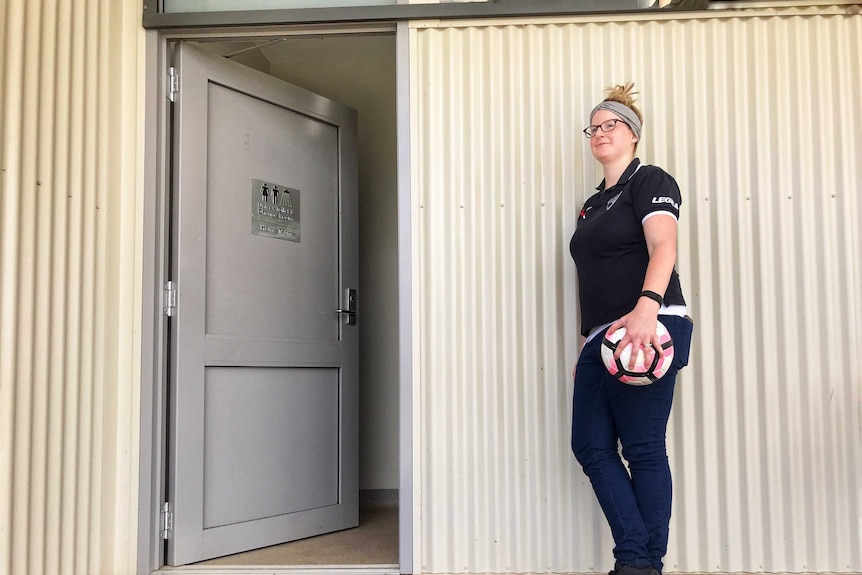 A woman in sports clothes stands next to a shed-like changeroom, holding a soccer ball.