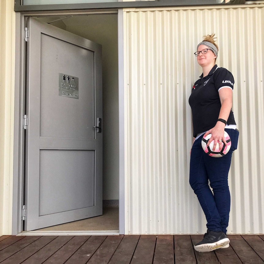 A woman in sports clothes stands next to a shed-like changeroom, holding a soccer ball.