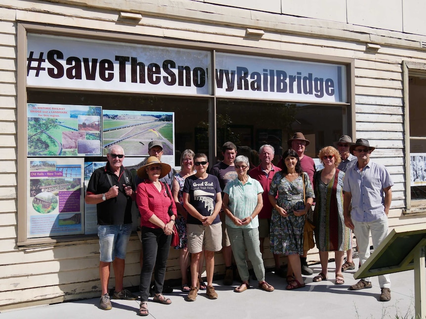 A group of people stand outside a shopfront featuring a large sign that reads #SaveTheSnowyRailBridge