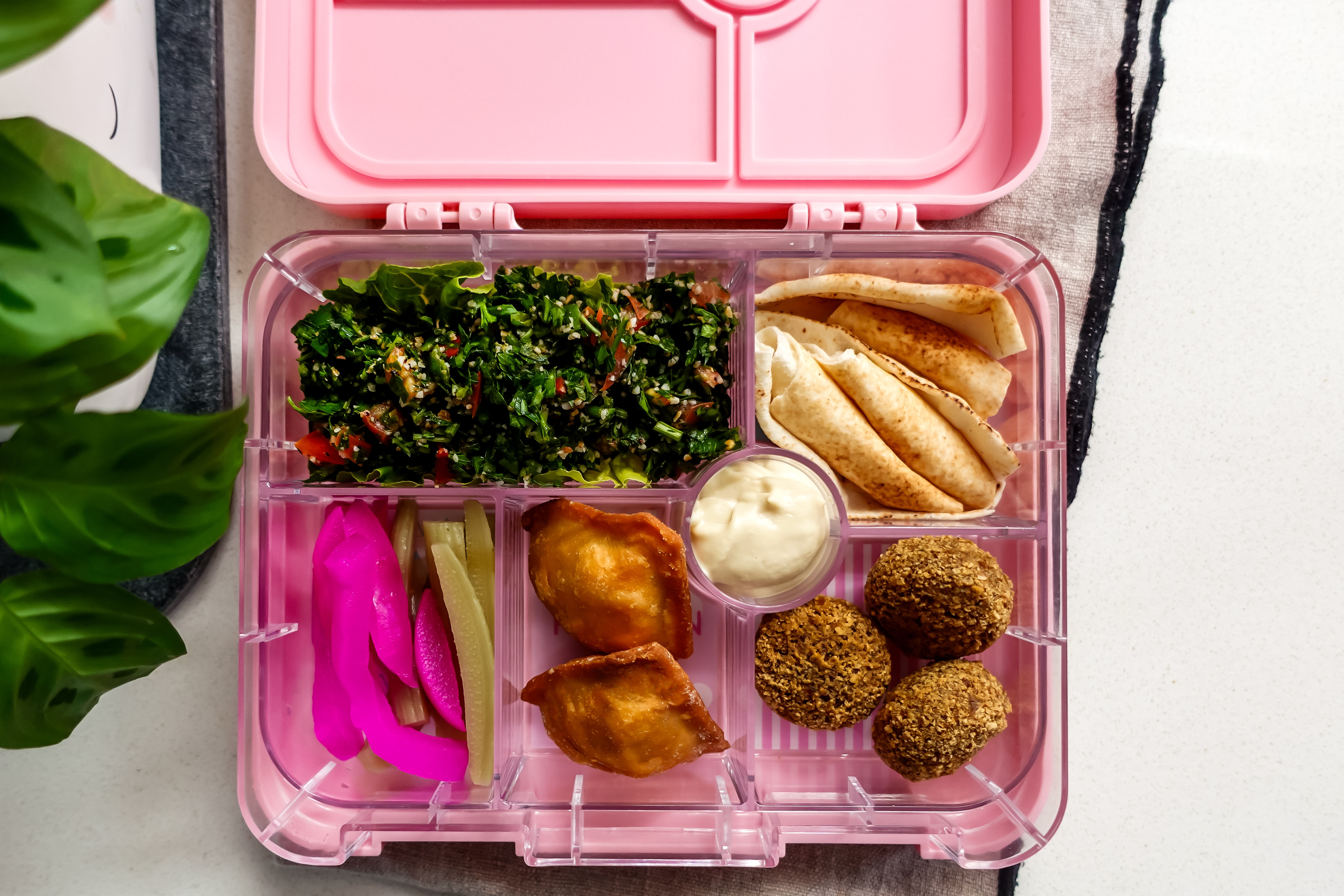 A pink lunchbox with sections with homemade tabouli, pickles, bread, hummus and falafel.