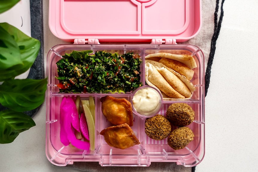 A pink lunchbox with sections with homemade tabouli, pickles, bread, hummus and falafel.