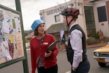 A woman and a man look at each other while wearing an ice cream tub and a spiky bicycle helmet on their heads.