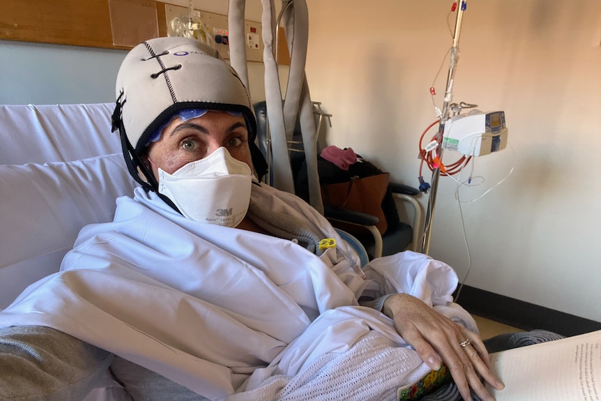 Nina Di Santo sits in a hospital bed wearing a facemask and a monitoring cap