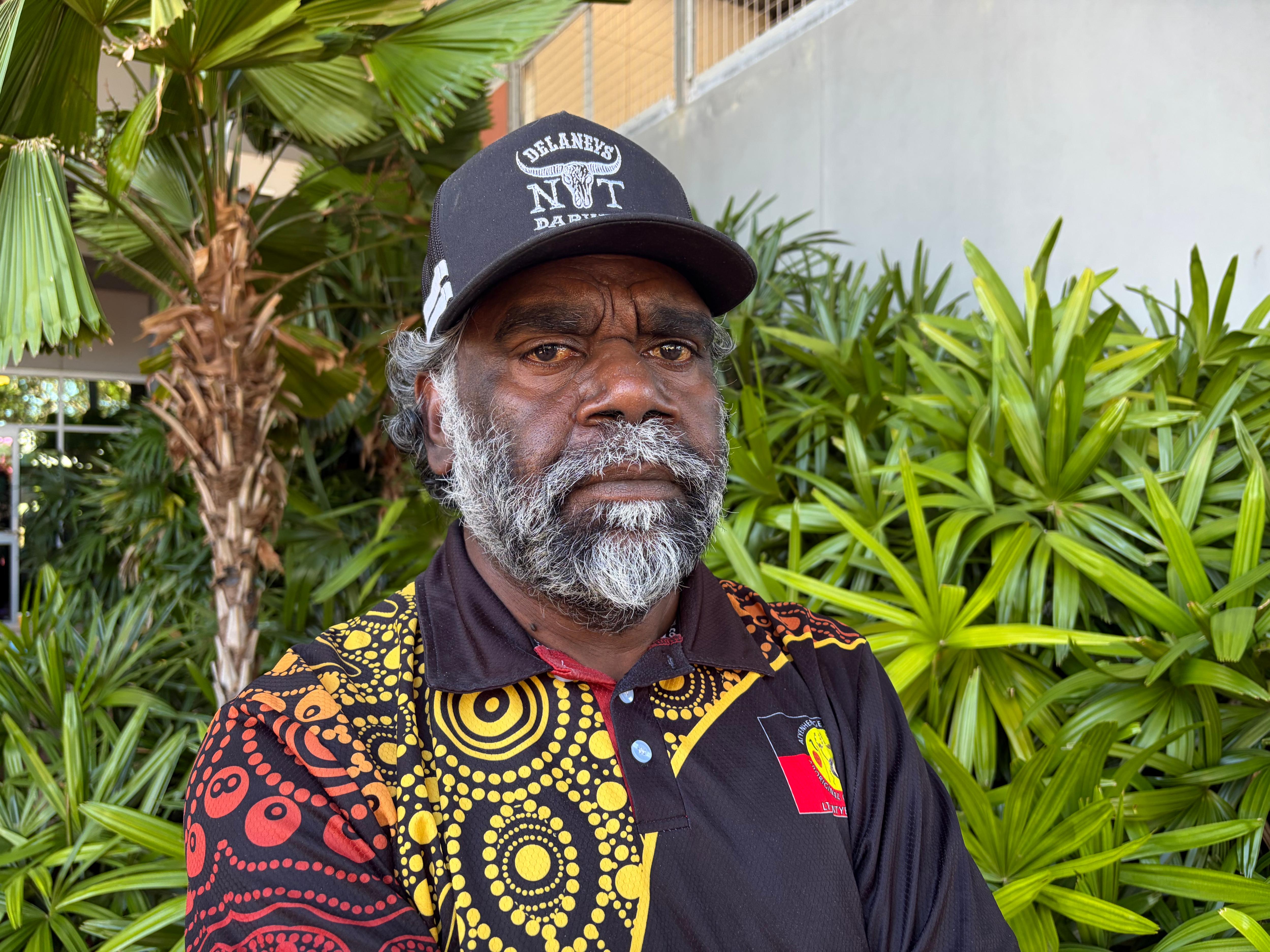 Hombre con camisa polo con estampado de arte indígena mirando a la cámara mientras está parado frente a plantas verdes 