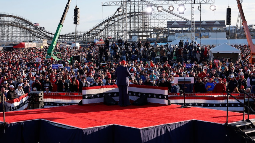 Trump on a red stage at a rally with a roller coaster behind