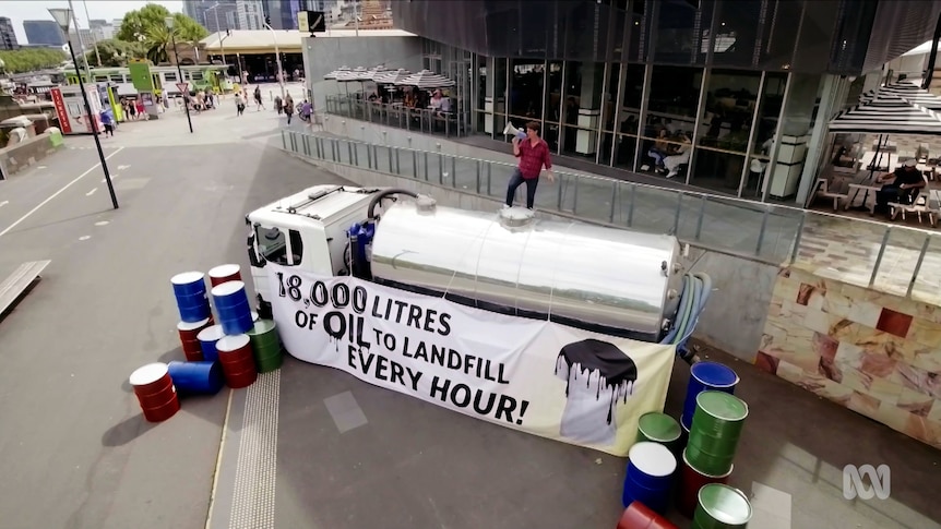 Craig Reucassel stands on an oil tanker, sign reads '18,000 litres of oil to landfill every hour!'