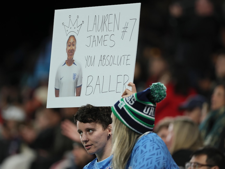 Two football fans stand in the grandstand, holding up a sign saying 'Lauren James #7 you absolute baller'. 