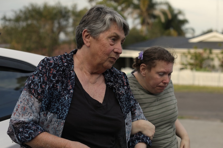 A woman with greying hair links arm her arm through her daughter's as they walk from a car. Behind them is a suburban street.