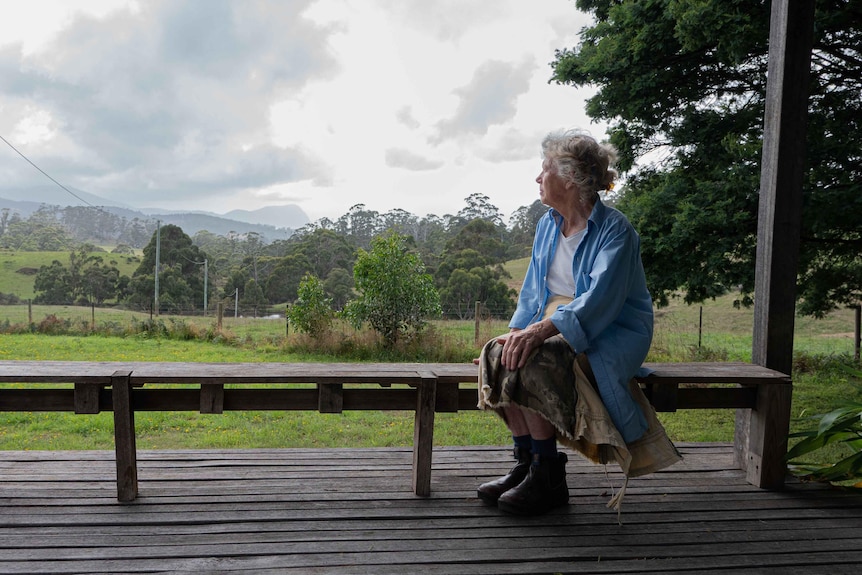 a woman sitting on a wooden bench looking out at a view of greens hills and a distant bluff