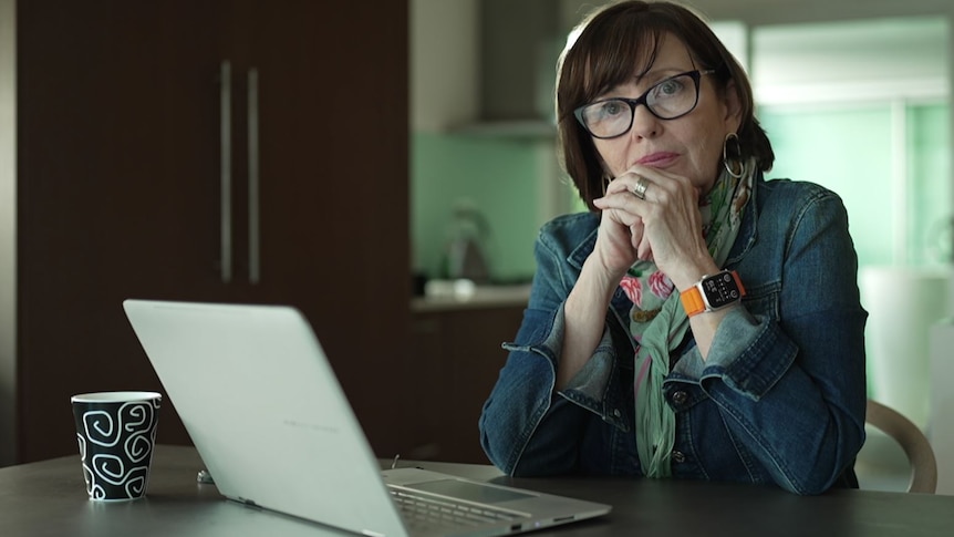 Woman with brown hair and a fringe wearing black-rim glasses sits looking serious at a laptop