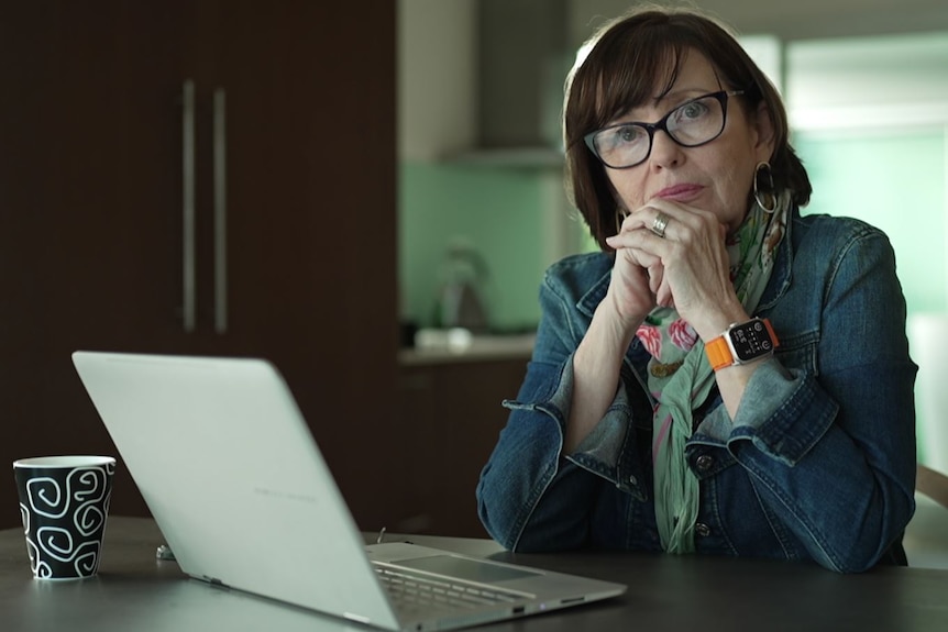 Woman with brown hair and a fringe wearing black-rim glasses sits looking serious at a laptop