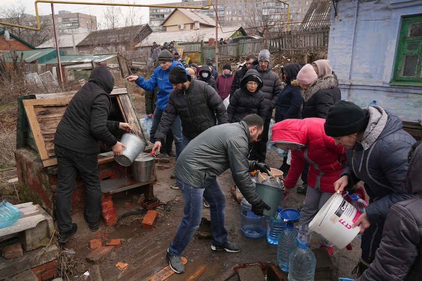 People in winter clothes using buckets to move water into large plastic water bottles.
