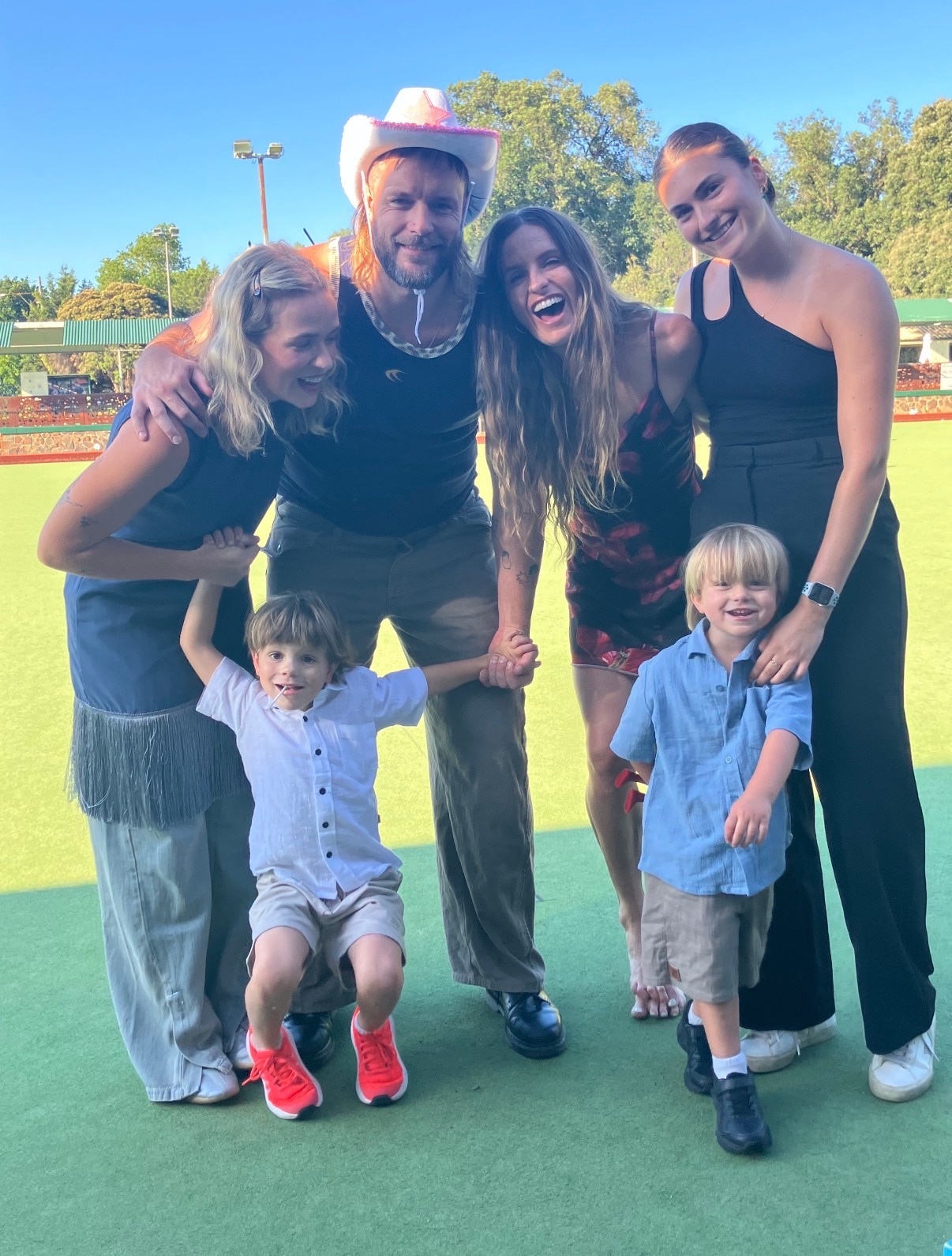 Um grupo de quatro adultos e duas crianças posam em um gramado verde, Mitch Brown usando um chapéu de cowboy rosa, todos sorrindo
