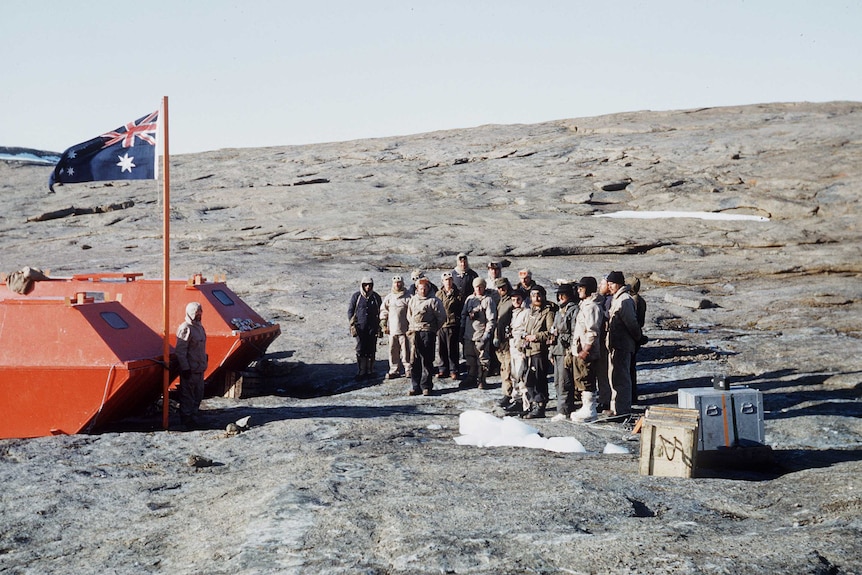 A group of men in thick winter clothes standing on a rocky slope in Antarctica, the Australian flag flying above them.