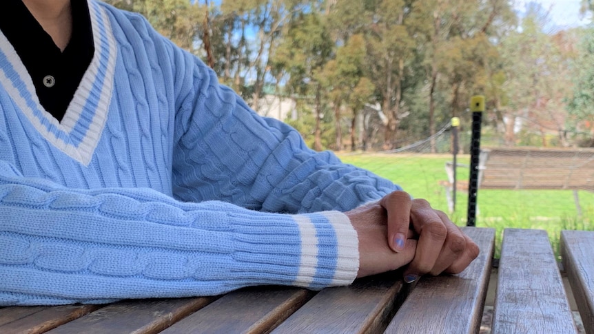 A woman's upper torso is shown as she sits on a park bench, with a clear blue sky behind her. Her face is not shown.