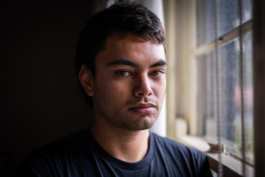 A young man standing by a window, looks directly into the camera. Half his face is in shadow.