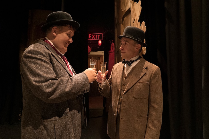Colour still of Steve Coogan and John C. Reilly clinking glasses backstage in 2018 film Stan and Ollie.