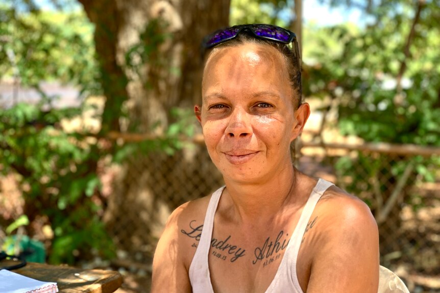 A woman with tatoos and a singlet smiles at the camera while sitting in a shady yard