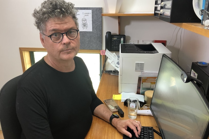 A middle-aged man with grey, curly hair and wearing a black T-shirt and glasses, sits at a desk with a keyboard and PC screen.