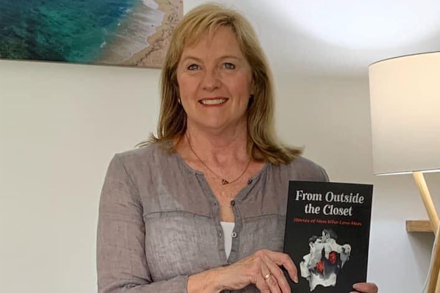 A woman with short sandy hair holds a book with a dark cover titled 'From Outside the Closet'