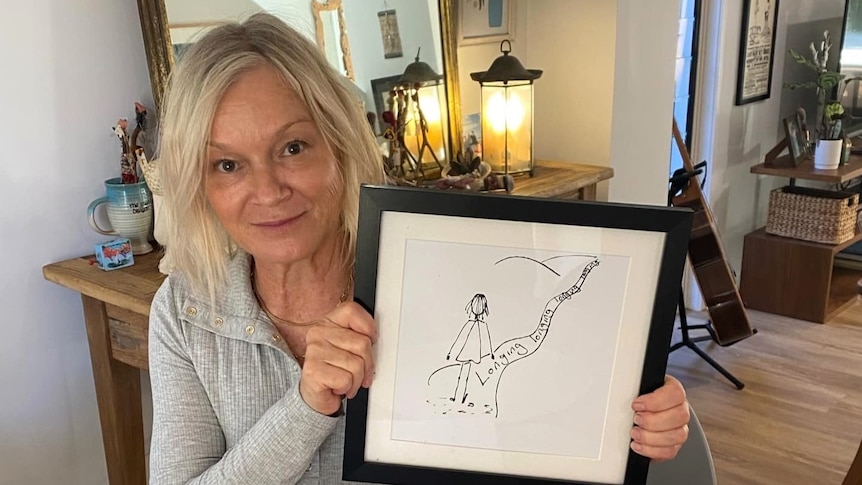 Woman holding up a framed drawing of a stick figure with the words, 'Longing, longing, longing'