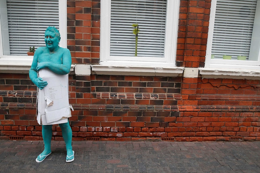 Man with a cardboard box wrapped around him walks around a building