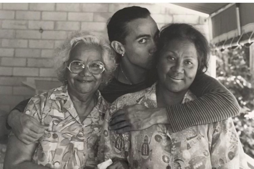 A black-and-white photograph of a younger man with his arms around two older women, whose arms are interlinked, all smiling.