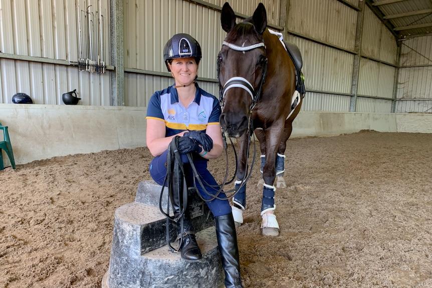 A woman in horse riding helmet sitting on a stool with a brown horse standing next to her