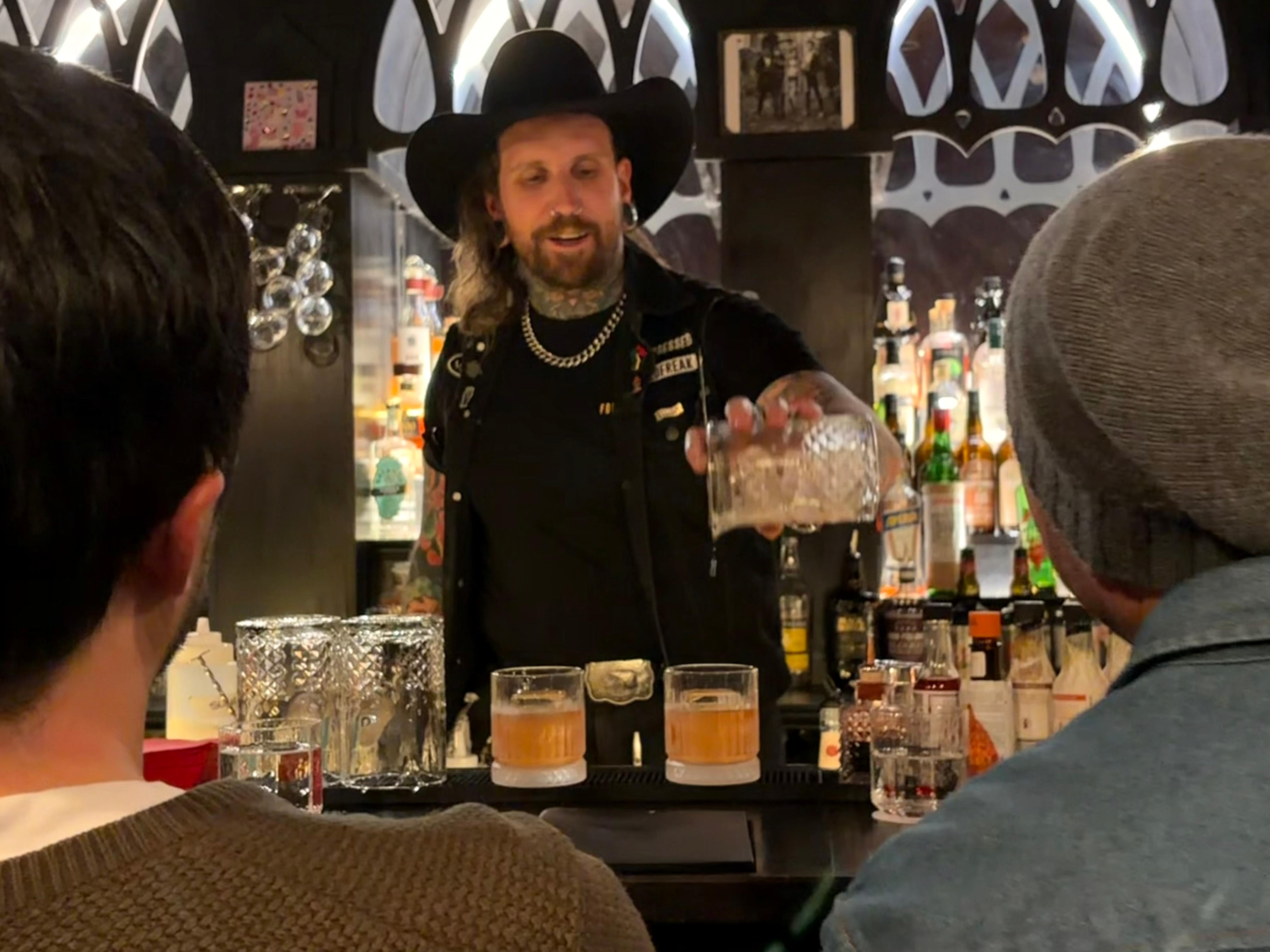 Man in dark bar, clothes and hat pouring drink into glasses between two people
