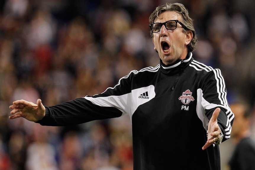 A middle-aged coach in glasses and black striped tracksuit top yells and separates arms on sideline of game with crowd behind.