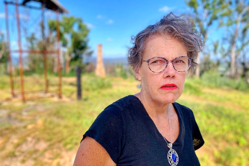 A grey-haired woman with glasses stands looking serious at an empty block of land.