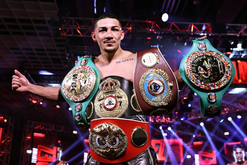 Teofimo Lopez holds three belts across his outstretched arms and wears two across his waist.