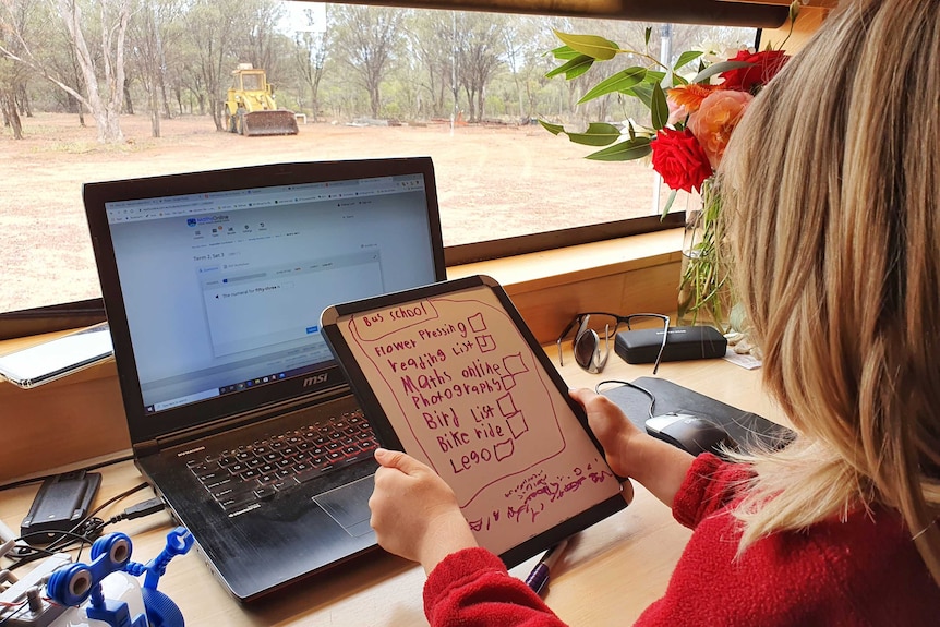 A child looks at a handheld whiteboard with a laptop on a table doing home-schooling in outback Queensland.