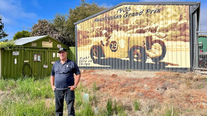A man stands in long grass in front of a shed painted with a large mural of a 1930s racing car.