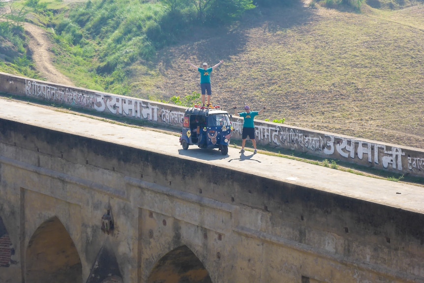 A man stands on a tuk-tuk on a large bridge with paddocks in the background.