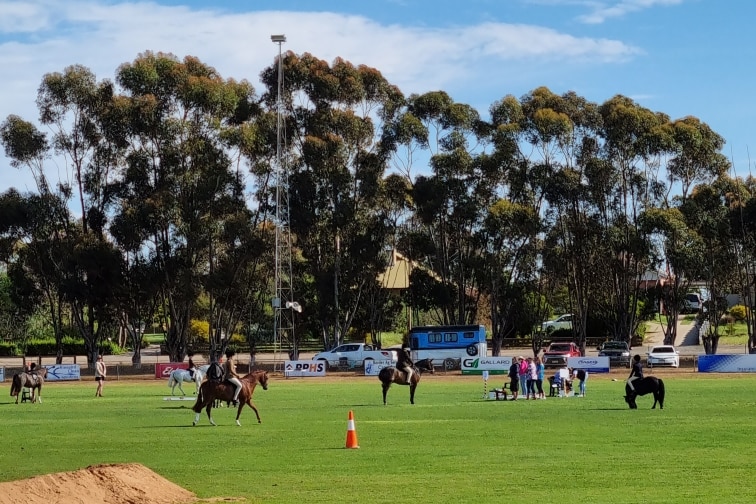 Horses with riders gallop in a field, with chairs in the foreground for spectators at the Pinnaroo Show