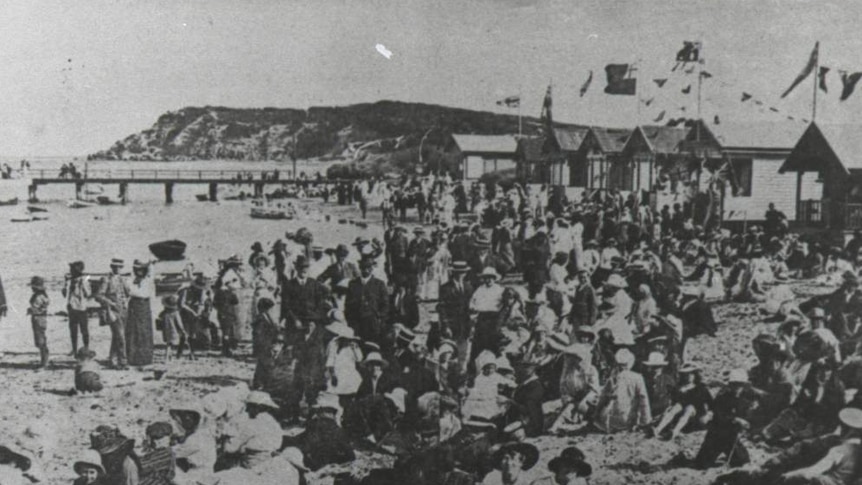 A black and white photo of hundreds of people  on a beach beside bathing boxes with a bridge in the background.