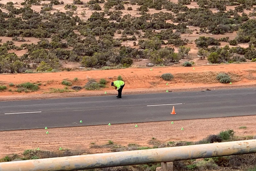 A police officer in a yellow fluoro jacket bends over looking at a road with orange cones on it