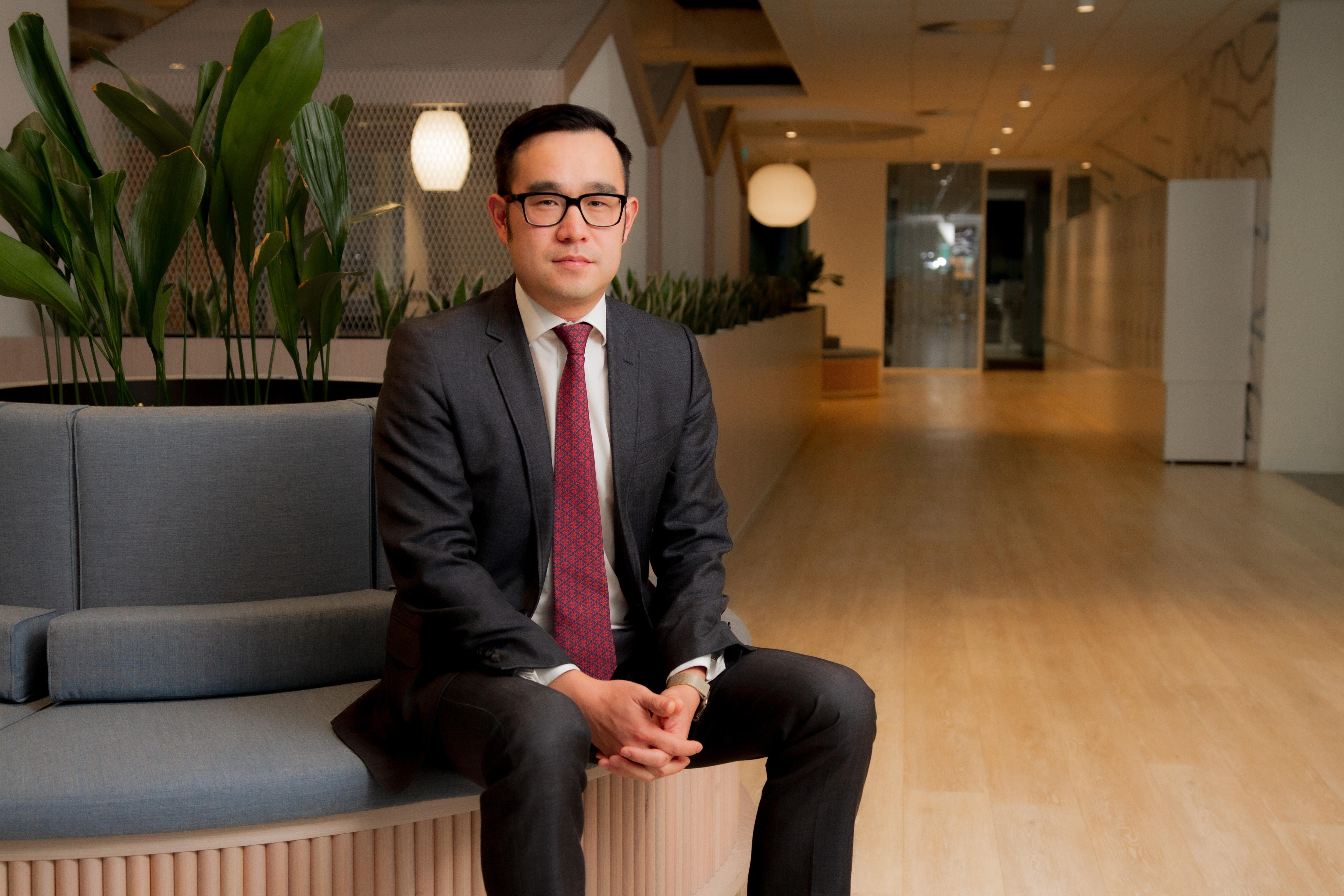 A bespectacled man with short, dark hair wears a dark suit as he sits in an expensive-looking corporate lobby.