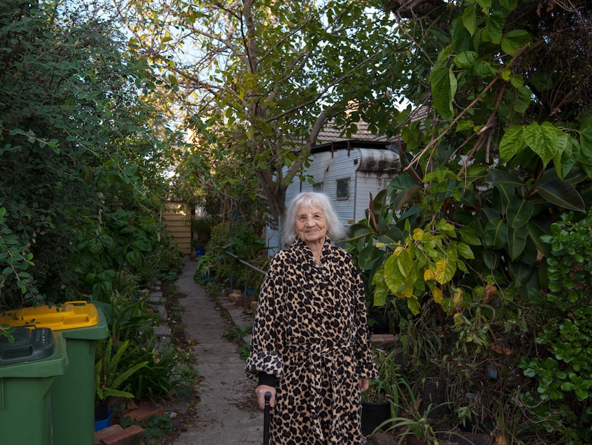 A woman in a leopard print dressing gown stands in a suburban yard, surrounded by lush foliage