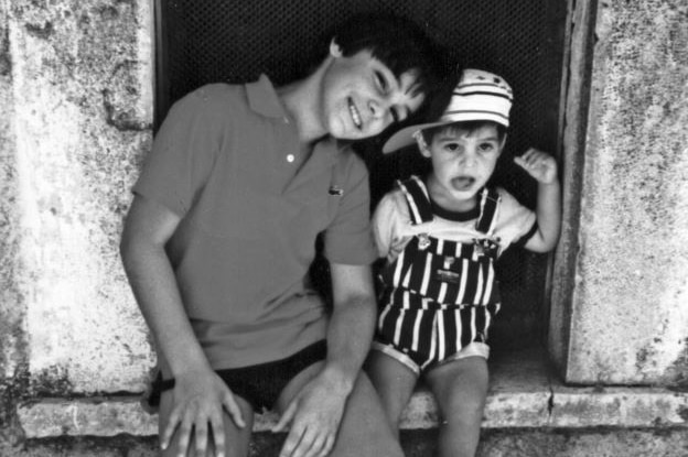 Black and white photo of a smiling teenage boy in t-shirt, shorts, long socks and sneakers seated next to toddler in overalls.