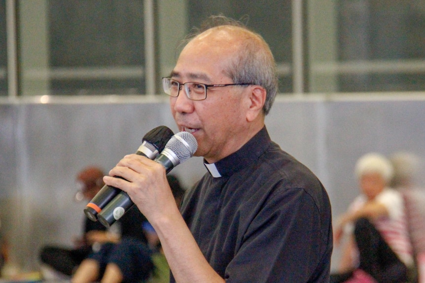 A man in a priest outfit holds two microphones