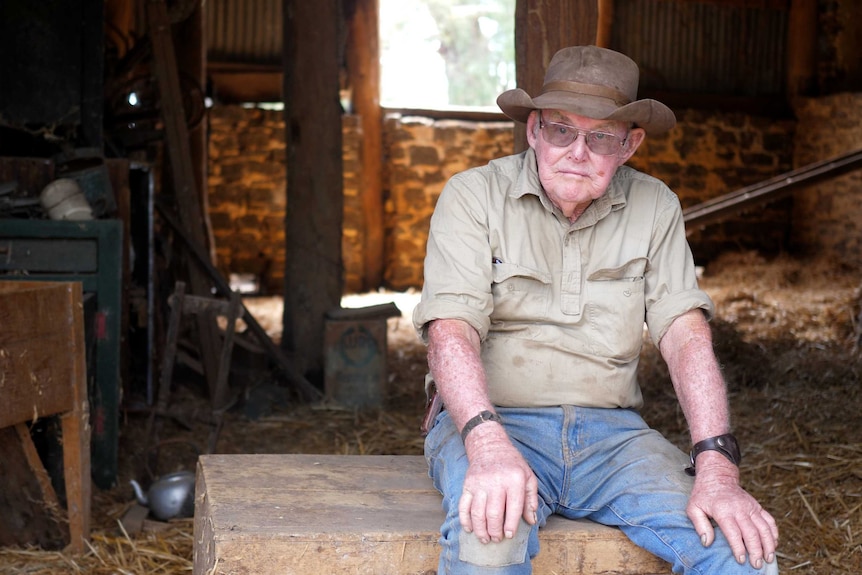An elderly farmer ina nbroad hat and glasses sits on an old trunk in an old hay shed.