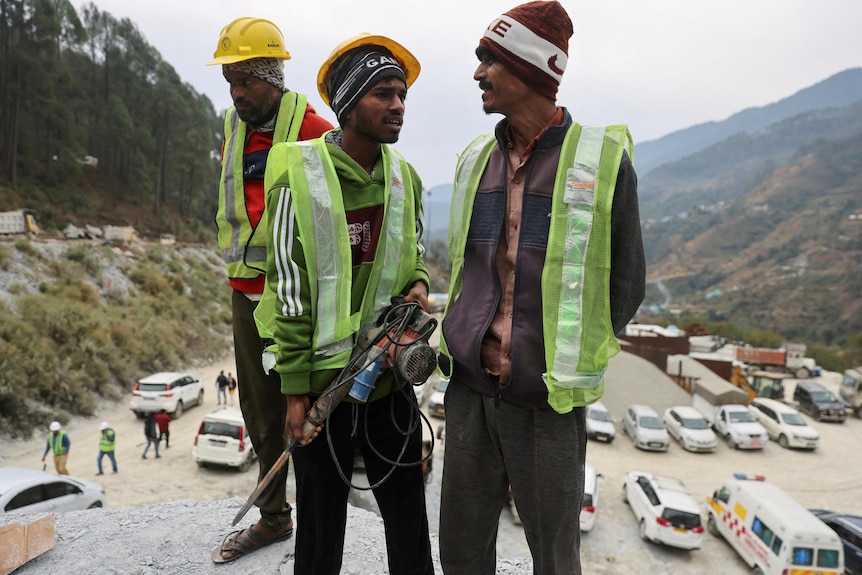 Three Indian men in hi-vis vests stand, one carrying a jackhammer. 