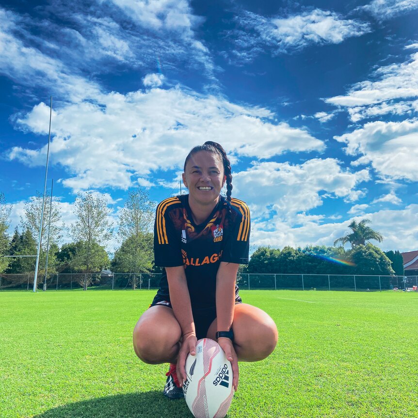 Crystal Kaua smiling in a Chiefs Manawa jersey squatting down on a rugby field leaning on a rugby ball on a sunny day