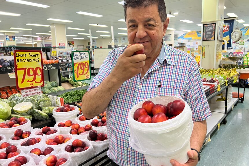 A dark-haired, middle-aged man holds a nectarine up to his mouth while holding a bucket of the fruit in his grocery shop.