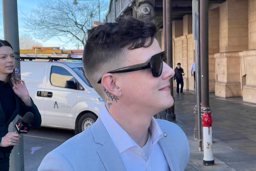 A man wearing sunglasses and a light blue jacket outside a stone building